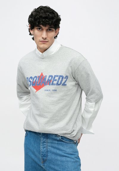 Pelēka sporta džemperis ar zilu un sarkanu rakstītu tekstu "DSQUARED2" un "SINCE 1995." Valkāts pār baltu kreklu ar apkakli. Klasiskas zilas džinsa bikses.