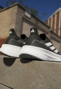 Baskets de sport noires et grises au design épuré, avec une semelle intermédiaire blanche rembourrée, une semelle extérieure texturée et trois rayures blanches sur les côtés.