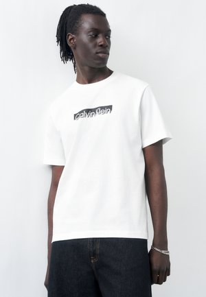 Mlad moški s temno poltjo in dredlji nosi belo majico Calvin Klein in temne kavbojke, gleda na stran ob enostavni svetli podlagi.