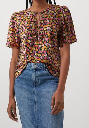 Vrouw draagt een korte mouwen blouse met bloemenprint in roze, oranje en geel, gecombineerd met een high-waisted blauwe denim rok.