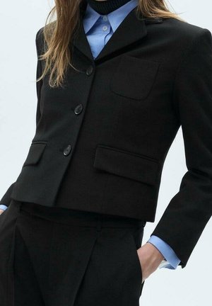 Frau trägt einen taillierten schwarzen Blazer mit Knöpfen, ein blaues Hemd mit Kragen und passende schwarze Hose, steht mit einer Hand in der Tasche.