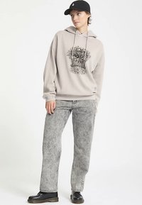 Sweat à capuche gris clair avec un motif frontal, associé à un jean gris délavé et des bottines noires. Le mannequin porte une casquette noire.