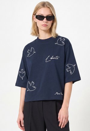 Blonde Frau mit schwarzen Sonnenbrillen und einem dunkelblauen Hemd mit weißen Vogelskizzen und dem darauf gedruckten Wort "Liberté".