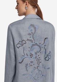 Ljusblåa linnejacka med intrikata broderade blommönster i ljusare nyanser. Noterbara urklippsdetaljer förbättrar texturen och mönstret.