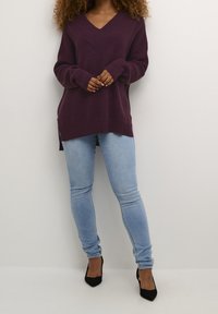 Burgundy stickad tröja med V-ringning, med ribbad textur och sidokilar, matchad med ljusblå tajta jeans och svarta högklackade skor.