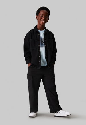 Camisa de mezclilla negra de botones, camiseta gráfica azul clara, pantalones cargo negros con bolsillos y zapatillas deportivas blancas. Fondo neutro.