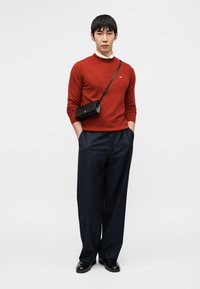 Κόκκινο πλεκτό πουλόβερ, λευκό γιακά, μπλε σκούρο παντελόνι με λεπτές ρίγες, μαύρη τσάντα χιαστί και μαύρα παπούτσια. Άνδρας στέκεται με τα χέρια στις τσέπες.