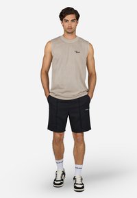 Ærmeløs beige bomulds skjorte med logo, parret med sorte shorts med sidesyninger. Modellen har sorte og hvide sneakers på med logosokker.