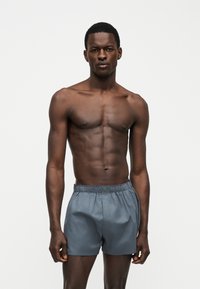 Homme torse nu à la musculature développée, debout face à l’objectif, portant un short gris à taille élastique sur un fond clair uni.