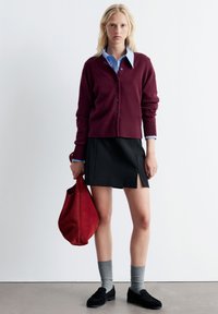 Burgunderfarbener Cardigan, hellblaues Hemd mit Kragen, schwarzer Rock mit seitlichem Schlitz, graue Socken, schwarze Slipper und eine rote Wildleder-Handtasche.