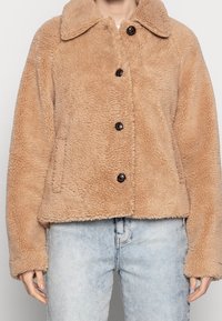 Giacca beige fluffy con bottoni e collo grande, indossata con jeans azzurri, che mostra il torso e le mani di una persona.