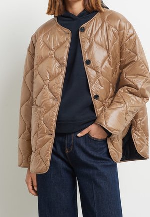 Le mannequin porte une veste matelassée beige sur un haut bleu marine et un jean foncé, une main dans une poche.