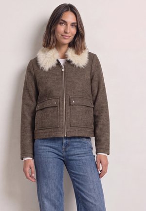 Femme portant une veste marron à fermeture éclair avec un grand col en fausse fourrure crème et un jean bleu, debout contre un mur clair uni.