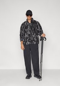 Veste floue à motif noir et gris avec une fermeture éclair, assortie à un pantalon sombre et une casquette noire. La personne tient un bâton de randonnée.
