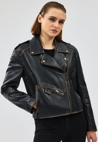 Giubbotto di pelle nera con un design aderente, zip asimmetrica e spalline. Presenta cuciture visibili e dettagli di cucitura in contrasto.