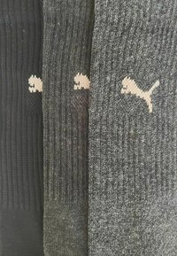 Drei Paar Socken: schwarz, dunkelgrau und hellgrau. Jede mit gerippter Textur und einem kleinen, tanfarbigen Puma-Logo an der Seite.