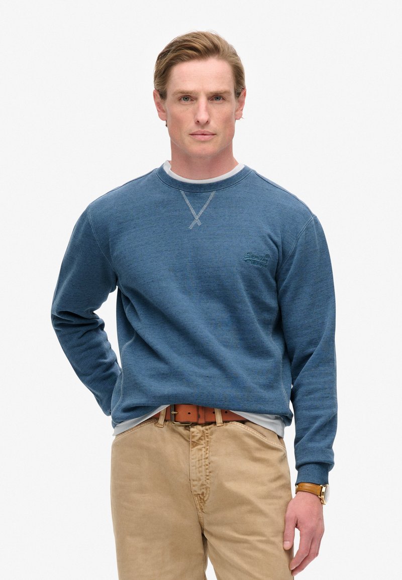 Sudadera azul de cuello redondo con mangas largas y un detalle de cruz bordado en el cuello, llevada sobre una camisa blanca, combinada con pantalones caqui.