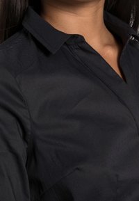 Zbliżenie na osobę w czarnej koszuli z kołnierzykiem, z widoczną linią dekoltu i obszarem ramion.