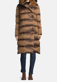 Braune Steppjacke mit Reißverschluss und Druckknopfverschluss, Kapuze und breiten horizontalen Steppnähten. Getragen zu dunklen Jeans und braunen Stiefeln.