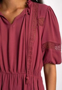 Vestido burdeos con escote en V, cuello volado, acentos de encaje en las mangas y cintura elástica; presenta una tela suave y texturada.