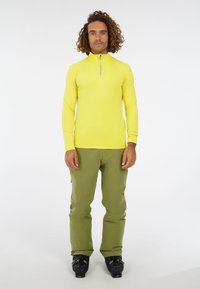 Man die staat in een felgele langarmige ritssluiting-top, olijfgroene broek en zwarte skibotten tegen een eenvoudige witte achtergrond.