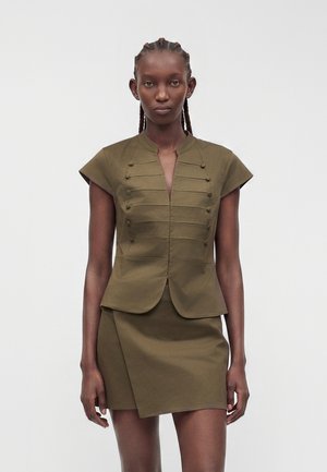 Femme aux cheveux tressés portant un haut olive à manches courtes avec des détails de boutons et une mini-jupe asymétrique assortie, debout devant un fond uni.
