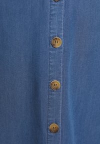 Ljusblå denimtyg med en vertikal rad av fem runda, ljusbruna knappar med två små hål i varje. Slät textur, avslappnad design.