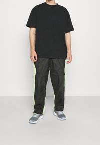 Černé oversize tričko kombinované s černými kalhotami s neonově zelenými bočními panely. Šedé tenisky dokončují outfit. Jednoduchý, moderní design.