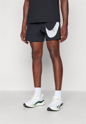 Black athletic shorts with a large white Nike swoosh logo, paired with white sneakers featuring green accents and white socks.
