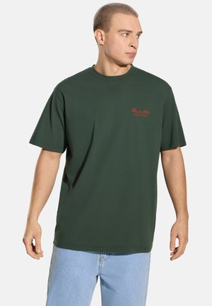 Zielony bawełniany T-shirt z krótkimi rękawami, z małym czerwonym haftowanym logo na piersi i luźnym krojem. Połączony z jasnoniebieskimi dżinsami.