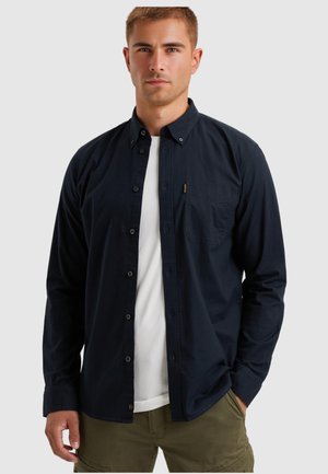 Uomo che indossa una camicia blu navy a bottone sopra una maglietta bianca con pantaloni verde oliva, in piedi contro uno sfondo grigio semplice.