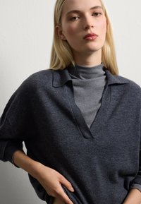 Maglione oversized grigio scuro a maglia con scollatura a V e maniche lunghe indossato sopra un dolcevita grigio. Texture liscia, design minimal, vestibilità rilassata.