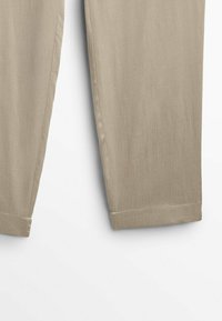 Dolna część beżowych spodni o prostym kroju z widoczną teksturą tkaniny i wykończonymi szwami na białym tle.