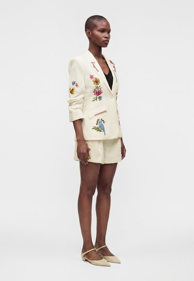 Femme debout en ensemble blazer et short crème avec broderies florales et oiseaux colorées, haut noir, chaussures plates neutres, fond blanc.