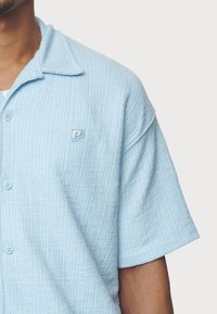 Vaaleansininen, teksturoitu lyhythihainen paita, jossa on kaulus, napillinen etuosa ja vasemmassa rinnassa kirjailtu logo. Pehmeä, resorikangas.