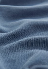 Puha, kék anyag texturált, bordázott mintával. Az anyag könnyűnek és enyhén gyűröttnek tűnik, hangsúlyozva annak hajlékony jellegét.