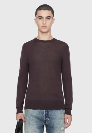 Ung mand med kort hår iført en mørkebrun langærmet trøje og lyseblå jeans, der holder en sort taske-rem i venstre hånd.
