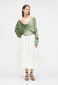 Γυναίκα που φοράει φαρδιά πράσινη σατέν μπλούζα, λευκή πλισέ φούστα μέσης μήκους και μαύρα σανδάλια με τακούνι, στέκεται με το ένα χέρι στη μέση.