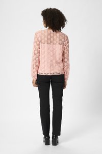 Haut en dentelle rose à manches longues, avec un motif floral et des détails ajourés, associé à un pantalon noir ajusté et des chaussures à talons.