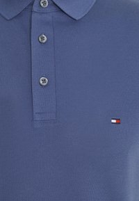 Polo bleu texturé avec trois boutons gris et un petit logo rectangulaire rouge, blanc et bleu marine sur la poitrine.