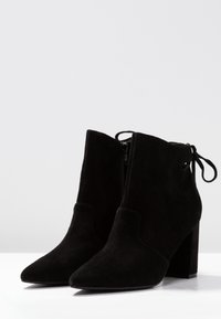 Botas de tobillo de ante negro con puntera afilada, cremallera lateral y un tacón ancho. Cuenta con un detalle de encaje en la parte posterior para el ajuste.