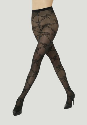 Gambe con calze nere trasparenti con motivo e scarpe nere con tacco alto incrociate davanti a uno sfondo semplice.