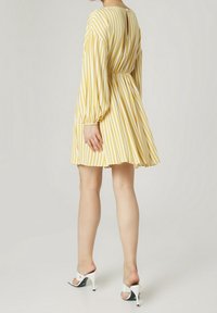 Žluté a bílé pruhované šaty s dlouhými rukávy, elastickým pasem a rozšířenou sukní; mají klíčovou dírku na zádech a jsou vyrobeny z jemné látky. Oblečeno s bílými lodičkami.