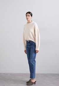 Suéter beige claro con mangas largas y sueltas, combinado con jeans de denim azul y zuecos trenzados marrones; diseño simple con textura suave.