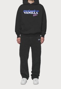 Schwarzer Kapuzenpullover mit großem blauem und pinkem Grafiktext auf der Vorderseite. Kombiniert mit schwarzer Hose und weißen Turnschuhen.