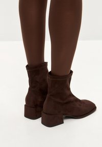 Bocanci din suede maro, până la gleznă, cu vârful pătrat și toc mic de tip bloc, având o margine simplu coaptă și o textură moale.