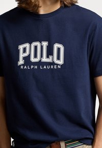 Námořnická modrá bavlněná tričko s bílou, tučnou grafikou "POLO" a textem "RALPH LAUREN" níže. Klasický design s kulatým výstřihem a krátkými rukávy.