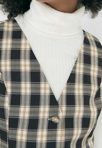 Gilet à carreaux dans des teintes de noir, beige et blanc. Présente un décolleté en V et une fermeture à un bouton. Superposé sur un col roulé blanc.