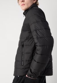 Svart pufferjakke med horisontal quilt, glidelåslomme på siden, høy krage og trykknapper ved ermer. Glatt tekstur.