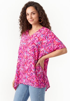 Kvinde med krøllet hår iført en lys pink mønstret bluse og lyseblå jeans, stående med den ene hånd på hoften og smilende.